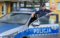 Policjant przy oznakowanym radiowozie i przed siedzibą Komendy Powiatowej Policji w Wieruszowie.