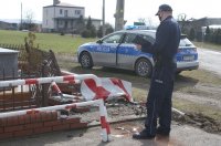 Na zdjęciu uszkodzone oznakowanie drogi, ogrodzenie kapliczki, umundurowany policjant i oznakowany radiowóz.