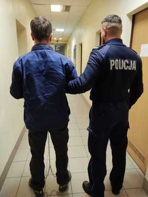 Na zdjęciu widoczny policjant, po jego lewej stronie mężczyzna w kajdankach zespolonych. Oboje stoją w pomieszczeniu dla osób zatrzymanych